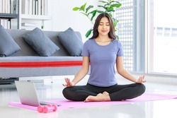 Asian young happy peaceful calm female model in casual sporty outfit sitting crossed legs in lotus position on yoga mat learning studying online meditation class via laptop computer in living room.