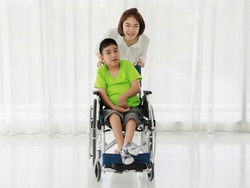Young positive Asian female caregiver rolling wheelchair with adorable smiling ethnic boy living with cerebral palsy disability in modern clinic
