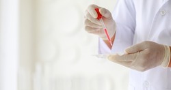 Closeup shot of scientist wears white lab coat and rubber gloves hands holding orange red reagent in glass dropper dropping into plastic plates for experiment in laboratory with blurred background.