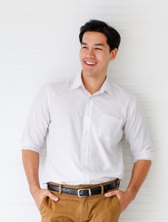 Portrait shot of young adult Asian man smiling in a formal white long sleeve shirt and brown pants with hand in pocket posturing looking at the side on white wall background in the studio.