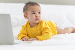 Portrait asian baby child look at camera do adorable and innocent near with laptop on bed in bedroom with bright light in the morning.