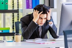 Closeup shot Asian young stressed depressed upset unhappy frowning face male businessman employee staff in formal suit sitting at working desk holding hands on head having migraine headache problem.