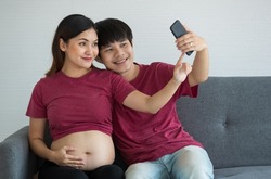 Happy smiley young couple dressed casually sitting on a couch together smiling and doing a selfie. They are expecting a healthy baby. Pregnancy and family concept.