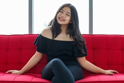 Front view portrait shot of a beautiful smiling young junior teenager in a black dress sitting on the couch and looking at the camera in the studio. Beautiful adolescent girl in prom party