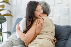 Asian cheerful young beautiful daughter sitting smiling hugging embracing showing affection love comforting bonding with old senior grey hair pensioner mother on cozy sofa in living room at home.