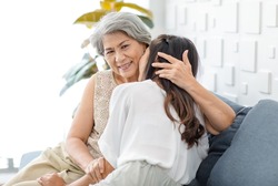 Asian cheerful young beautiful daughter sitting smiling hugging embracing showing affection love comforting bonding with old senior grey hair pensioner mother on cozy sofa in living room at home.