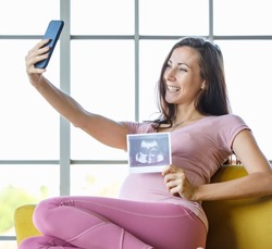 Millennial Caucasian young beautiful happy cheerful healthy big belly tummy pregnancy mother sit on sofa video call conversation via smartphone holding showing ultrasound sonogram Xray film picture.