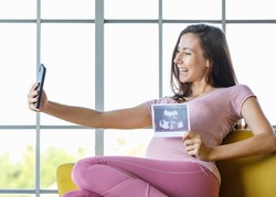 Millennial Caucasian young beautiful happy cheerful healthy big belly tummy pregnancy mother sit on sofa video call conversation via smartphone holding showing ultrasound sonogram Xray film picture.