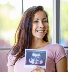 Portrait closeup shot of Millennial Caucasian young beautiful happy cheerful healthy pregnancy mother smiling look at camera holding showing ultrasound sonogram Xray film picture of unborn child.