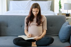 Caucasian millennial young happy female prenatal pregnant mother in casual brown pregnancy sitting on cozy sofa smiling holding showing ultrasound Xray scan picture in living room at home