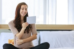 Caucasian millennial young happy female prenatal pregnant mother in casual brown pregnancy sitting on bed smiling holding showing ultrasound Xray scan picture in bedroom at home