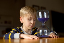 handsome blond boy sits at the table at home and considers an hourglass. Concepts of home schooling and education