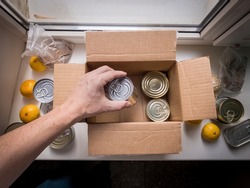 Needy man is unpacking package of donated volunteer food. Unpacking Cardboard Box of donations with products: canned food, cereals, citrus. Concept of food delivery during the coronavirus pandemic.