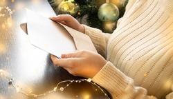 a girl in a sweater sits next to a Christmas tree and reads a letter.