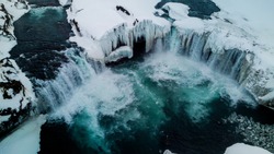 Aerial photo of a wide but not tall waterfall in Iceland. The waterfall is partly frozen and the landscape is covered in snow and ice. The drone took the photo from above.