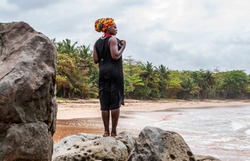 African woman stands on cliffs by the beach in Axim. Has headdress in traditional colors. Photo taken in Axim Ghana West Africa.