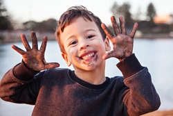 A head and shoulders photo of a handsome, smiling, dark haired, brown eyed little boy excitedly showing off the dirt on his face and hands after playing outdoors.