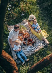 mom in a yellow dress and straw hat and dad in light clothes are sitting with their two young children under a tree on the grass on a picnic on a sunny day in summer top view