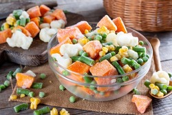 Mix of different frozen vegetables in a glass bowl on wooden table. Frozen food