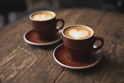 Two beautiful burgundy cups of cappuccino with trendy heart shape latte art. Summer loft cafe background. Urban space. Drinking coffee on the terrace.