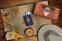 Top view of traveling gadgets, vintage map, magnify glass, hat and airplane model on the wood table background. On center, official passport of Antigua and Barbuda and your flag.