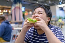 Obese boy eats a hamburger in a food court in a shopping center. junk food and unhealthy food concept