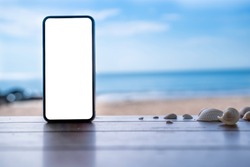 stock photo phone in the beach, black smartphone show white screen placed on wooden table with shells. Sky summer,beach and sea in background with copy space , blank for text ads and graphic design.