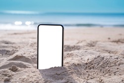 Smart phone on tropical sand beach with bokeh sun light, wave texture and blue sky background. White screen display  mock up for text, ads design. technology and travel nature concept with copy space.