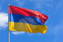 The flag of Armenia is a rectangular banner consisting of horizontal red, blue and orange stripes on a background of blue sky during the summer day.