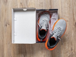 New sneakers in the open box on the floor. Pair of gray mesh fabric trainers with grooved orange sole in a cardboard box. Modern textile sneakers for sport and active lifestyle. Open new shoes box.