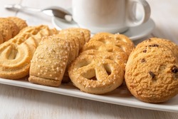 Assorted crispy shortbread biscuits on a rectangular plate and white tea cup. Set of danish butter cookies and coffee cup close-up. Breakfast with baked pastry. Sweet food concept. Front view.