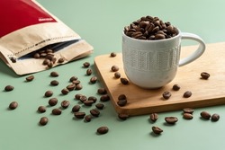 Roasted coffee beans in a small white cup on a pastel green background. Whole coffee beans spilled out of a brown paper bag. Organic coffee beans for making tasty strong beverage concept. Front view.