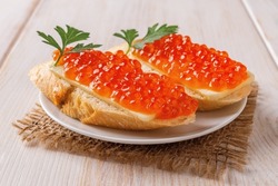 Two sandwiches with red caviar on a saucer over wooden table. Gourmet appetizer of trout caviar on a french baguette slice with butter. Salted salmon roe for fish delicacy concept. Front view.