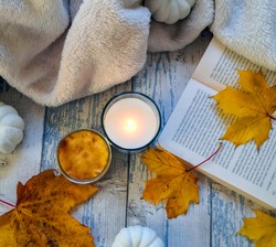 Autumnal composition on blue wooden surface with lighted candle, open book, blanket, white faux pumpkins and yellow maple leaves. Top view, from above, flat lay. Relaxing at home and hygge concept. 