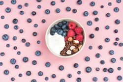 Concept of healthy breakfast.  Colorful bowl full of yoghurt, chocolate granola, raspberry, nuts and blueberry. Pink background with blueberry pattern