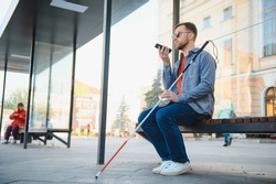 Young blind man with smartphone sitting on bench in park in city, calling
