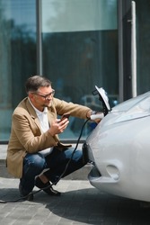 Hansome bearded man sitting near his new modern electric car and holding plug of the charger, while car is charging at the charging station.