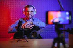 A young stylish guy with a beard wearing casual or music teacher playing guitar in front of smartphone camera. Online guitar training or vocal lesson.
