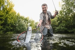Man with fishing rod, fisherman men in river water outdoor. Catching trout fish in net. Summer fishing hobby.