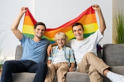 Lgbt family, gay couple with adopted son - Lgbtq parents with their kid having fun at home, family watching tv and holding lgbt rainbow flag
