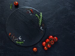 Dark food background with empty stone round plate,fresh rosemary,fresh cherry tomatoes,dry chilli peppers and sea salt on black background.Top view with copy space.