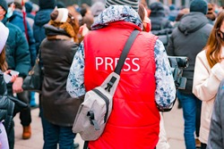 Correspondent in a red jacket with text “Press” and camera working in crowd. Protest. Strike. City. Outdoor. Job. Professional. Occupation. Specialist. News. Media. Broadcast. TV