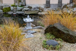 Rocks and yellow bushes near marble stone sculptural lantern on shore of pond with Japanese koi carp fishes. Autumn landscape design in eastern garden style. Fall season tranquil scene.