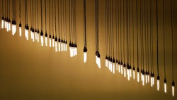 Rows of hanging pendant lights on long wire strings with yellow brown background. Glowing light bulbs hanging in diagonal row on blurred backdrop with copy space. Industrial style lamps.