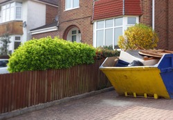 Industrial yellow rubbish skip on driveway. Selective focus on full metal bin with space to add text on surrounding background of green bush, wooden fence, houses. Renovate, moving, clearance concept.