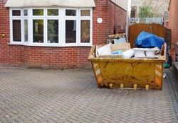 Full industrial rubbish bin on driveway in front of brick house with space to add text for background use, on side of heavy skip, pavement floor. Renovate, removal, recycle & home clearance concept.