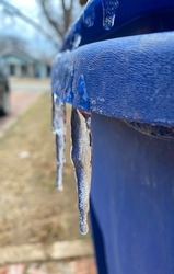 Many small icicles have frozen on blue trash can in the street in the winter.
