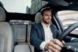 Success in motion. Handsome young man in full suit smiling while driving a car