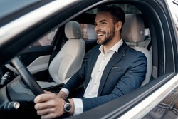 Man of style and status. Handsome young man in full suit smiling while driving a car