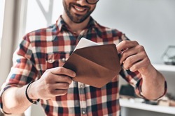 Receiving greeting card. Close up of young man opening envelope and smiling while standing indoors